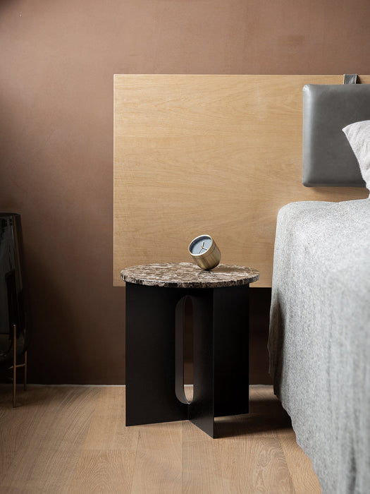 Androgyne Side Table with a black base and brown marble top, styled as a bedside table next to a bed with a wood headboard.