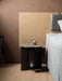 Androgyne Side Table with a black base and brown marble top, styled as a bedside table next to a bed with a wood headboard.