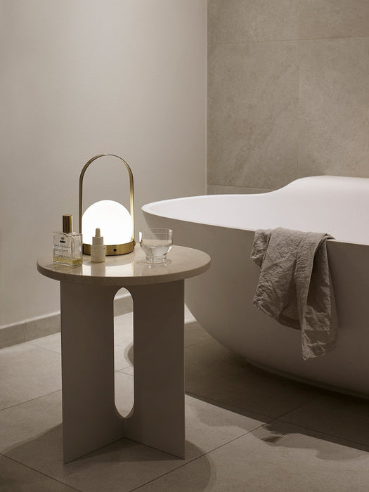 The ivory Androgyne Side Table with a glowing Carrie Lamp on top, placed next to a modern white bathtub in a calm bathroom.