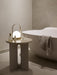 The ivory Androgyne Side Table with a glowing Carrie Lamp on top, placed next to a modern white bathtub in a calm bathroom.