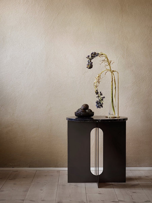 The black Androgyne Side Table with a marble top, featuring a central cutout, styled with vase and bowl against a beige wall.