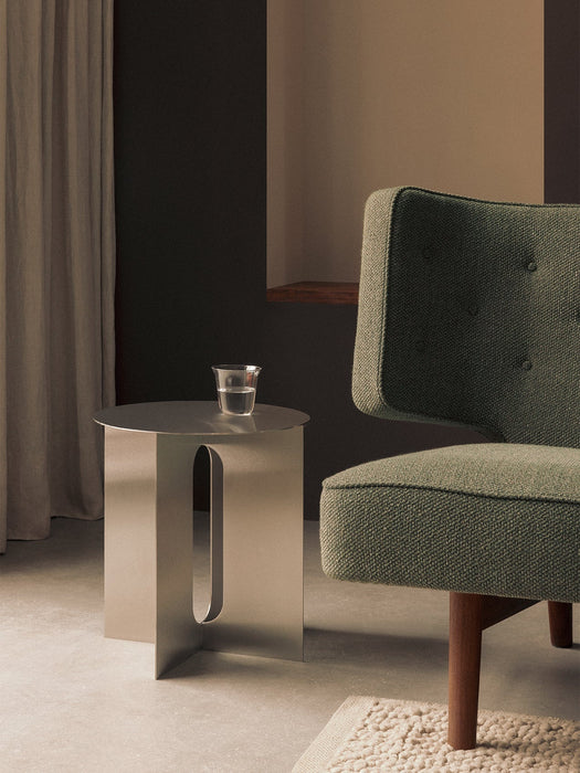 Androgyne Side Table in steel with a glass of water on it, styled next to a green armchair in a minimalist interior.