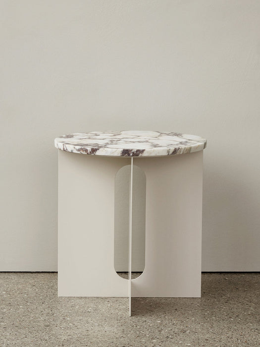 Androgyne Side Table with an ivory base and a veined marble top, standing on a terrazzo floor against a light grey wall.