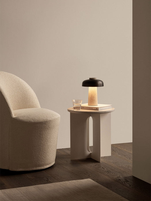 A cream Tearoom Club Chair next to an ivory Androgyne Side Table with a lit Reverse Table Lamp, a book and a glass on top.