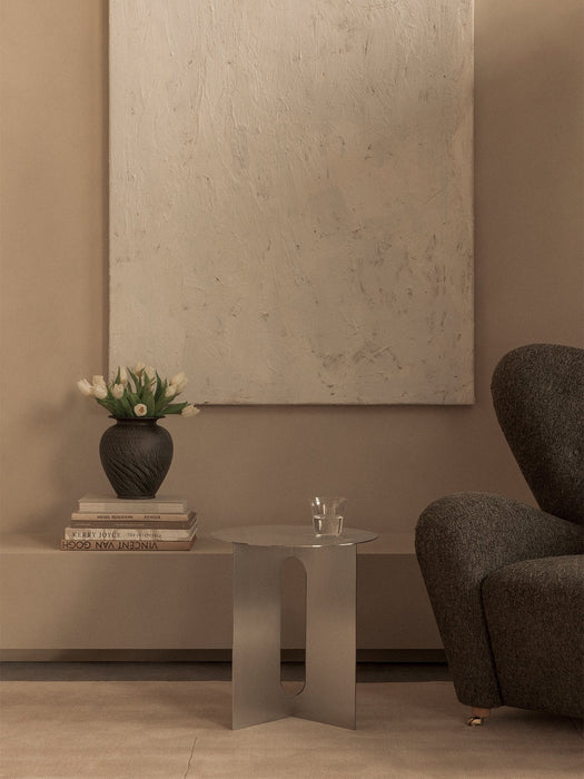 A steel Androgyne Side Table stands by a grey The Tired Man armchair in a beige room with a textured painting on the wall.