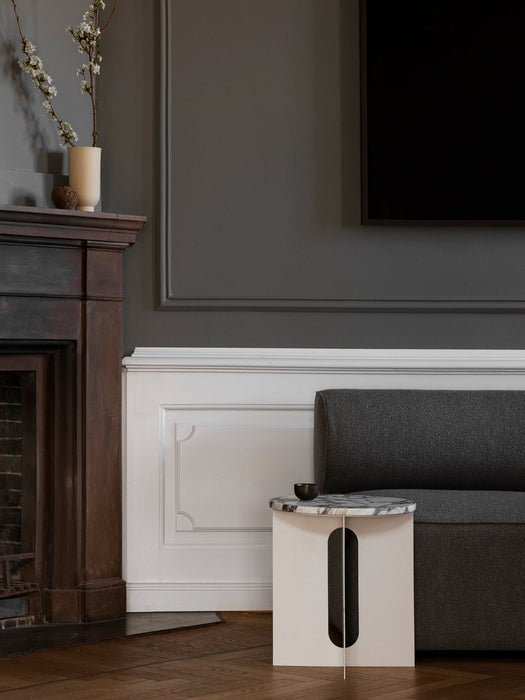 The Androgyne Side Table with an ivory base and marble top styled in a classic living room next to a grey sofa and fireplace.