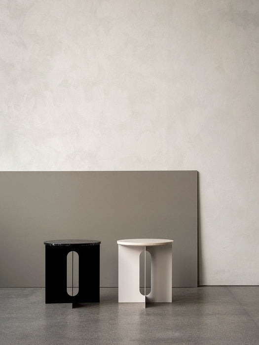 The Androgyne Side Table shown in black with a dark marble top and in ivory, standing side-by-side in a minimalist room.
