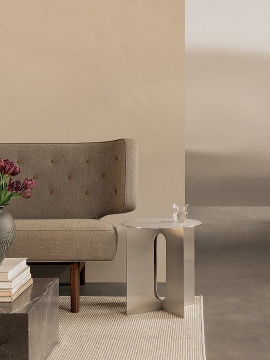 The stainless steel Androgyne Side Table with a glass on top, placed next to a brown button-tufted sofa in a neutral room.