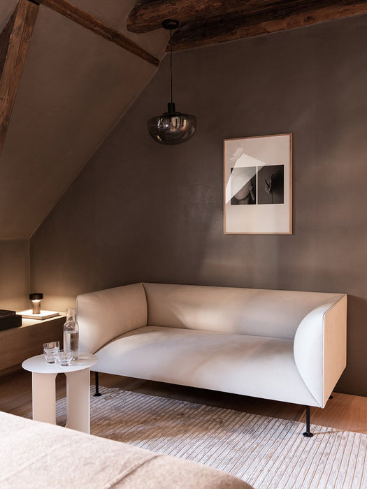 The white Androgyne Side Table sits beside a cream sofa in a dimly lit, modern room with a pendant lamp and framed artwork.