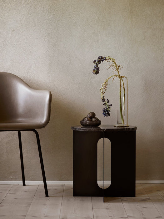 A black Androgyne Side Table with a marble top sits beside a taupe Harbour Chair. A Stem Vase with flowers is on the table.