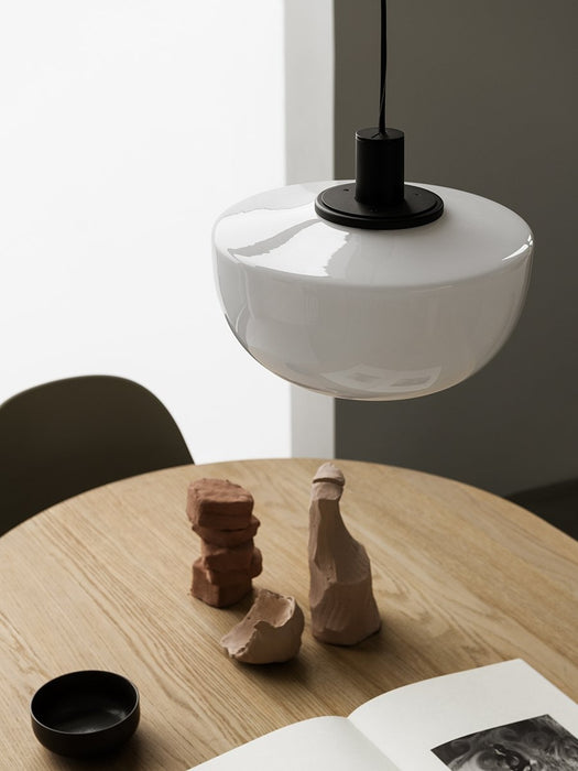 A close-up of the Bank Pendant with an opal glass shade and black fitting, hanging over a wooden table with clay sculptures.