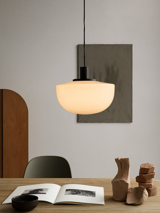 The Bank Pendant light with an opal glass shade glows over a wooden table with an open book and clay sculptures.