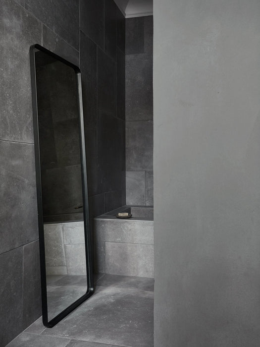 The tall, black-framed Norm Floor Mirror by Norm Architects leaning against a dark grey, stone-tiled wall in a bathroom.