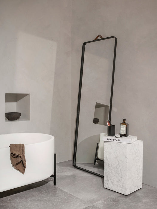 The Norm Floor Mirror leans next to a marble Plinth in a minimalist bathroom with a white freestanding tub.