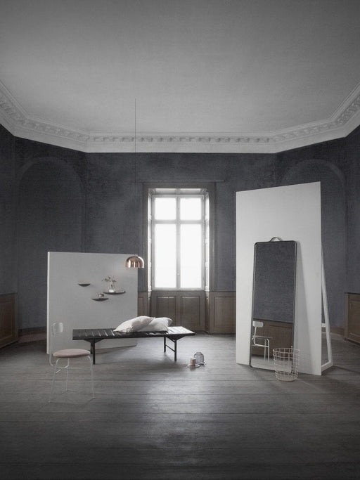The tall, black-framed Norm Floor Mirror stands in a minimalist room with dark walls, a daybed, and a hanging copper lamp.
