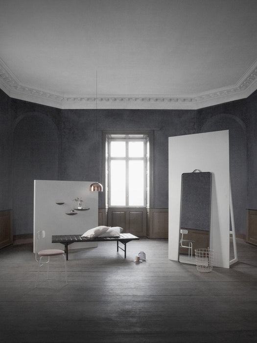 The tall, black-framed Norm Floor Mirror stands in a minimalist room with dark walls, a daybed, and a hanging copper lamp.