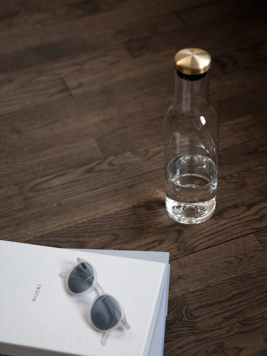A clear glass water bottle with a brass lid stands on a dark wood floor beside a white book with a pair of sunglasses on it.