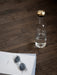 A clear glass water bottle with a brass lid stands on a dark wood floor beside a white book with a pair of sunglasses on it.