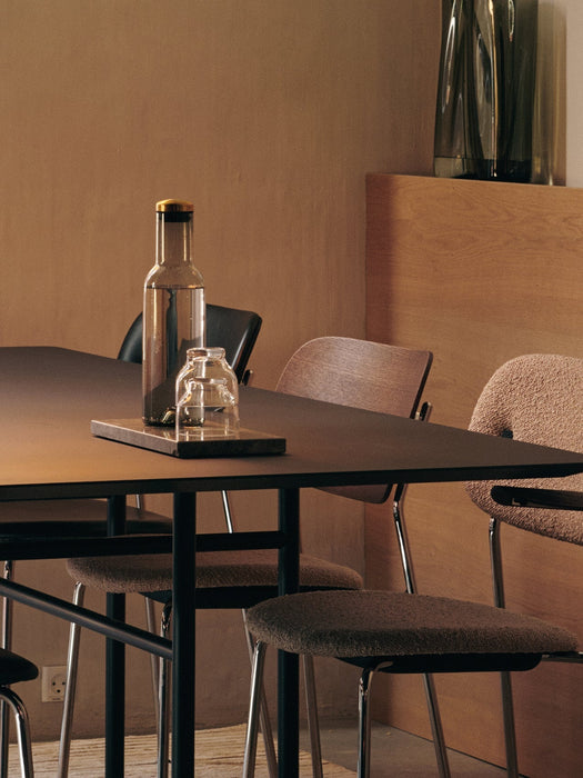 A warmly lit dining scene featuring a glass water carafe on a dark table, with minimalist wooden and upholstered chairs.