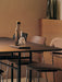 A warmly lit dining scene featuring a glass water carafe on a dark table, with minimalist wooden and upholstered chairs.