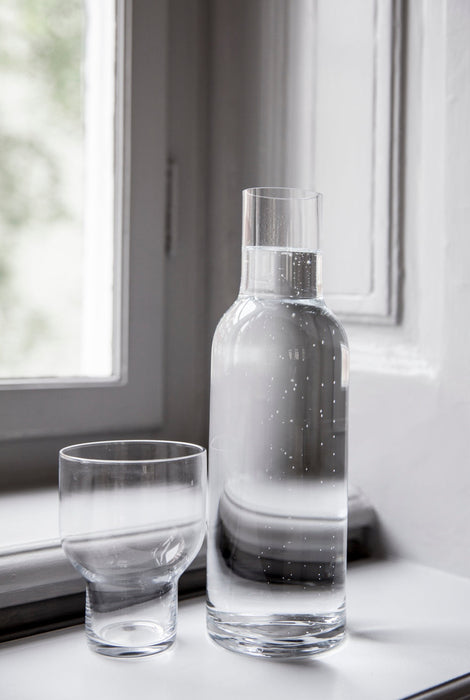 A tall, clear glass Stackable Bottle Carafe with bubbly water, beside a matching empty glass on a sunlit windowsill.