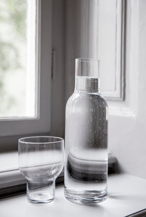 A tall, clear glass Stackable Bottle Carafe with bubbly water, beside a matching empty glass on a sunlit windowsill.