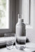 A tall, clear glass Stackable Bottle Carafe with bubbly water, beside a matching empty glass on a sunlit windowsill.