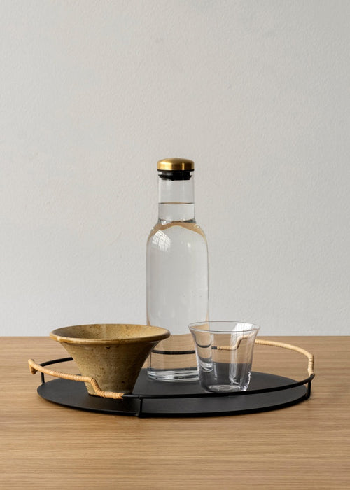 A black Balcony Serving Tray on a wooden table, holding a Bottle Carafe, a Strandgade Drinking Glass, and a Triptych Bowl.