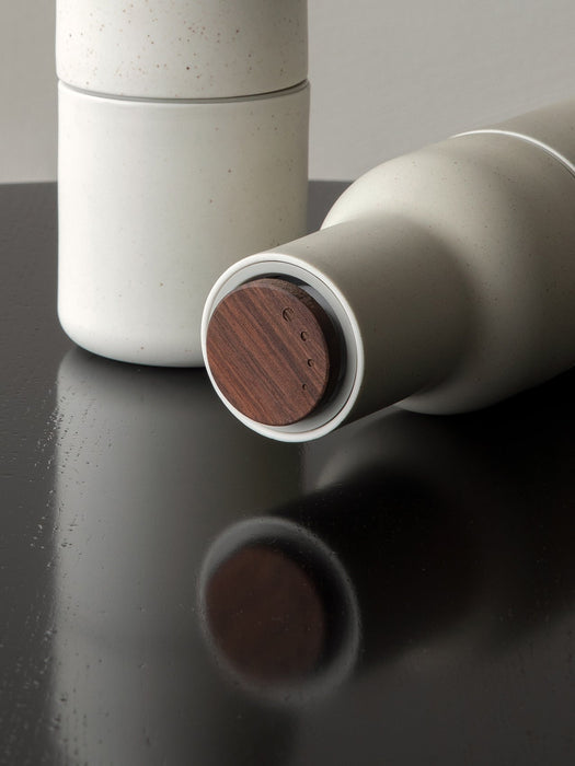 Close-up of a speckled off-white ceramic Bottle Grinder with a wooden base, lying on its side on a reflective dark surface.