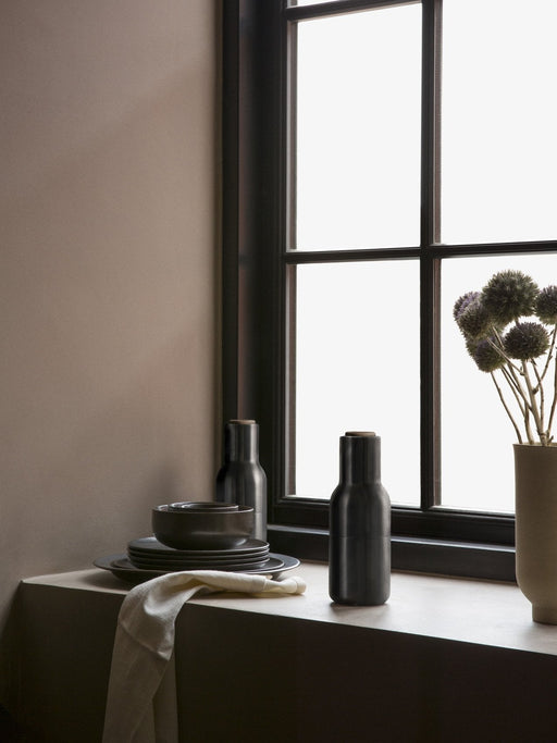 Two black Bottle Grinders sit on a stone windowsill next to a stack of dark grey plates, a napkin, and a vase of flowers.