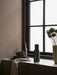 Two black Bottle Grinders sit on a stone windowsill next to a stack of dark grey plates, a napkin, and a vase of flowers.