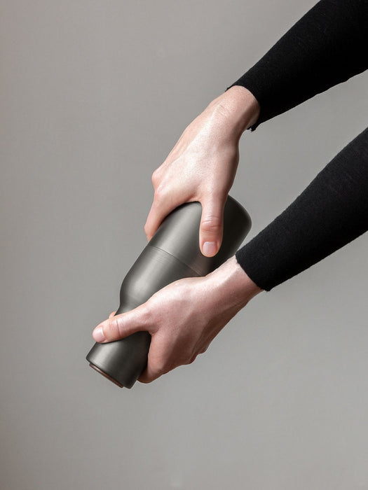 Close-up of two hands in black sleeves twisting the dark grey, matte Bottle Grinder from Audo Copenhagen on grey background.