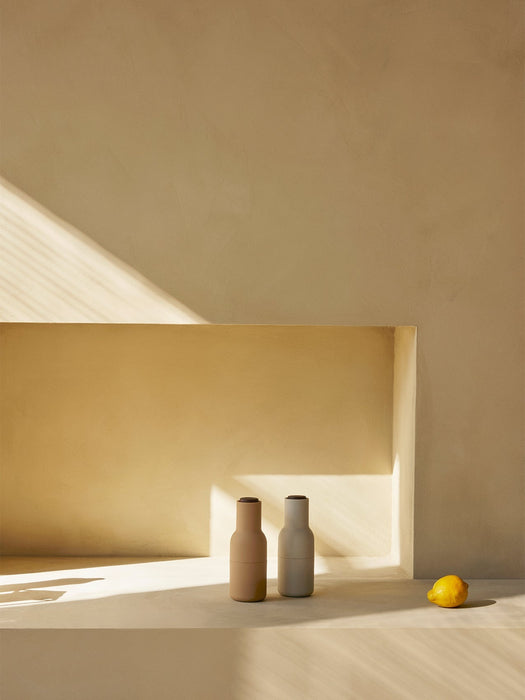 Two Audo Bottle Grinders, one beige and one light grey, on a stone surface in a sunlit, sandy-coloured niche next to a lemon.