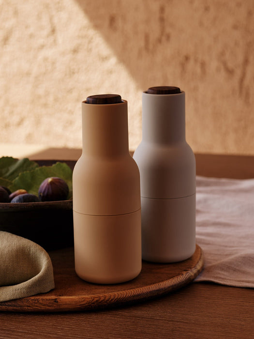 A lifestyle shot of two Bottle Grinders in sand and light grey with dark wooden lids, set on a wooden tray with fresh figs.