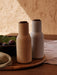 A lifestyle shot of two Bottle Grinders in sand and light grey with dark wooden lids, set on a wooden tray with fresh figs.