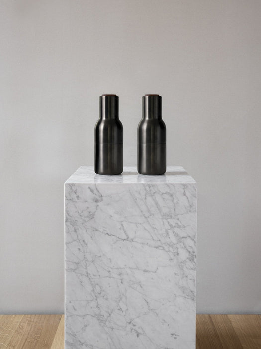 A pair of black Audo CPH Bottle Grinders displayed on a white marble plinth against a neutral grey wall and wooden floor.