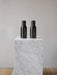 A pair of black Audo CPH Bottle Grinders displayed on a white marble plinth against a neutral grey wall and wooden floor.