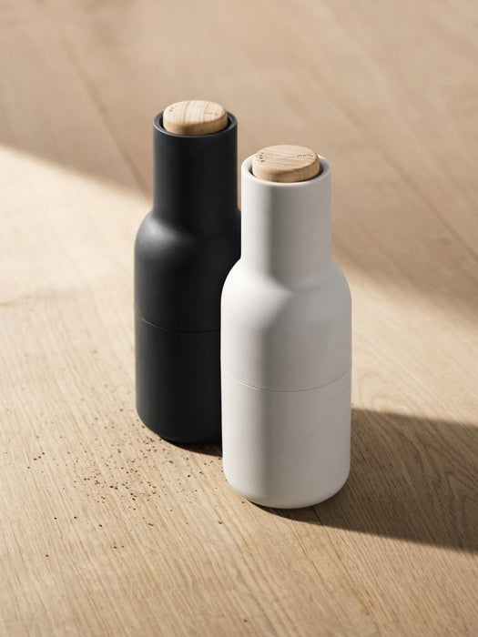 A black and light grey Bottle Grinder with wooden tops on a light wood surface, with freshly ground pepper scattered.