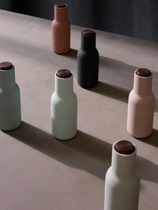 Six Audo Copenhagen Bottle Grinders in muted pastel and dark colours with wooden tops, arranged on a neutral-toned surface.