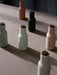 Six Audo Copenhagen Bottle Grinders in muted pastel and dark colours with wooden tops, arranged on a neutral-toned surface.