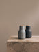 Two Audo Bottle Grinders, one light grey and one dark grey, standing on the corner of a beige marble plinth against a wall.