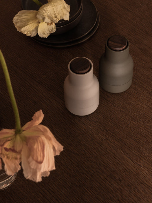 High-angle shot of two Audo Bottle Grinders, light grey and dark green, on a dark wood table with flowers and plates.