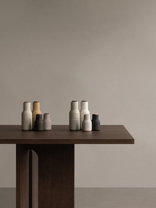 A collection of Audo Bottle Grinders in muted, earthy tones on the corner of a dark wood Androgyne Dining Table.