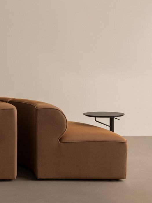 The black, round-topped Branch Side Table placed next to the arm of a brown Eave Seamline sofa against a beige wall.