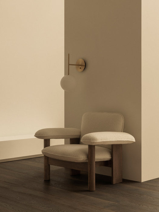 A Brasilia Lounge Chair with a low back in light textile sits on a dark wood floor next to a TR Bulb Suspended Wall Lamp.