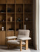 A Brasilia Lounge Chair in light bouclé fabric and oak frame sits on a Houkime Rug before a bookcase with a Torso Lamp.
