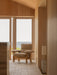 A beige Brasilia Lounge Chair with a light wood frame sits in a wood-panelled room, looking out of a window onto the sea.