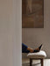 A person resting their feet, clad in black slippers, on the light-coloured, textured Brasilia Ottoman with dark wooden legs.