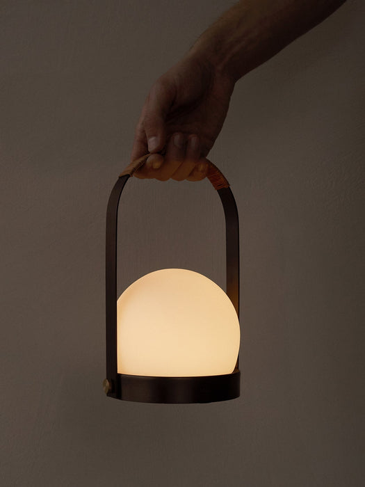 A hand holds the portable, illuminated Carrie Table Lamp. The bronzed brass frame with a leather handle holds a glowing orb.