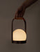 A hand holds the portable, illuminated Carrie Table Lamp. The bronzed brass frame with a leather handle holds a glowing orb.